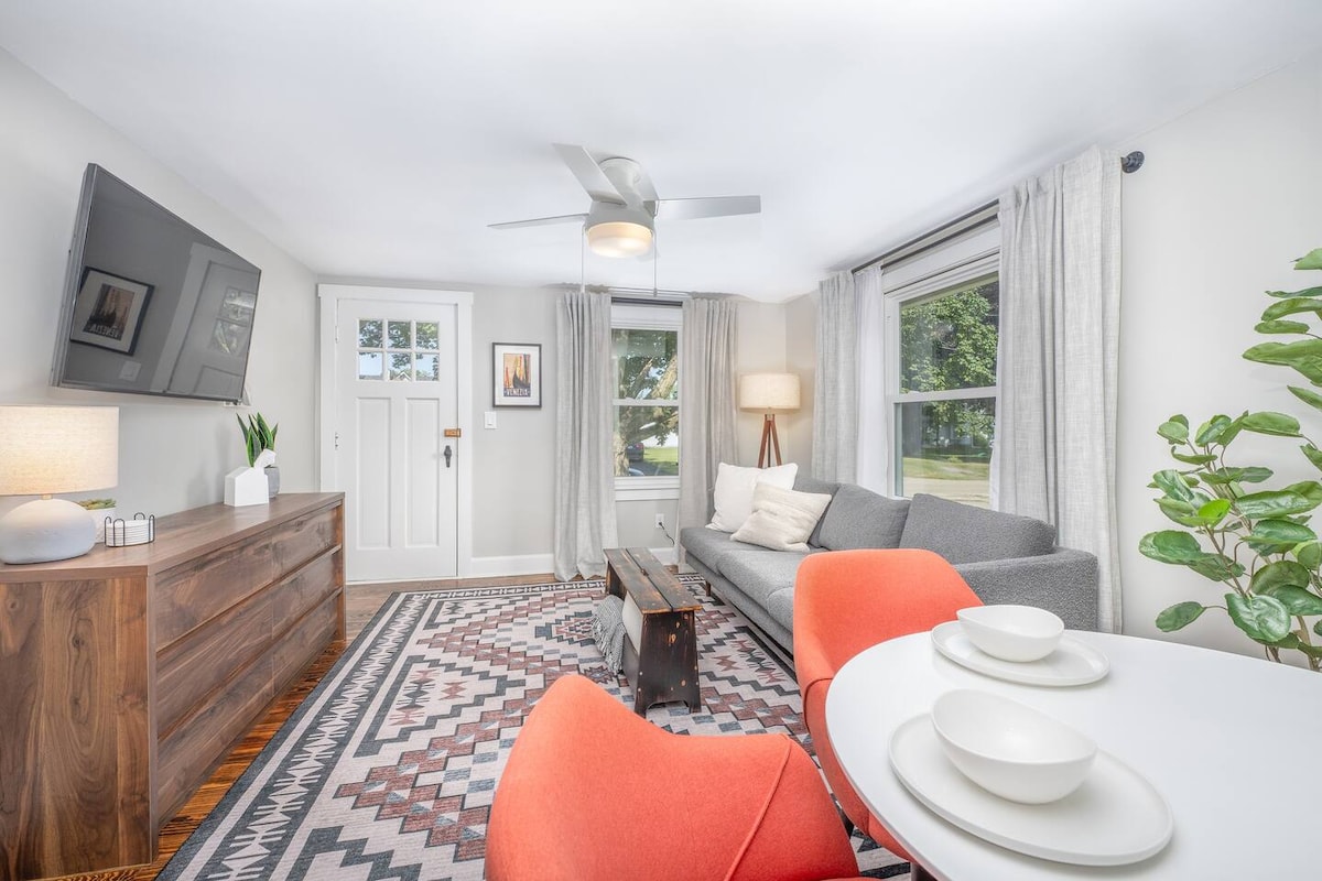 A light-filled living area features a modern sofa and a sleek coffee table, accompanied by bright chairs at a dining table set for two. Natural light pours in through large windows, while decorative elements enhance the cozy ambiance, creating a welcoming space.