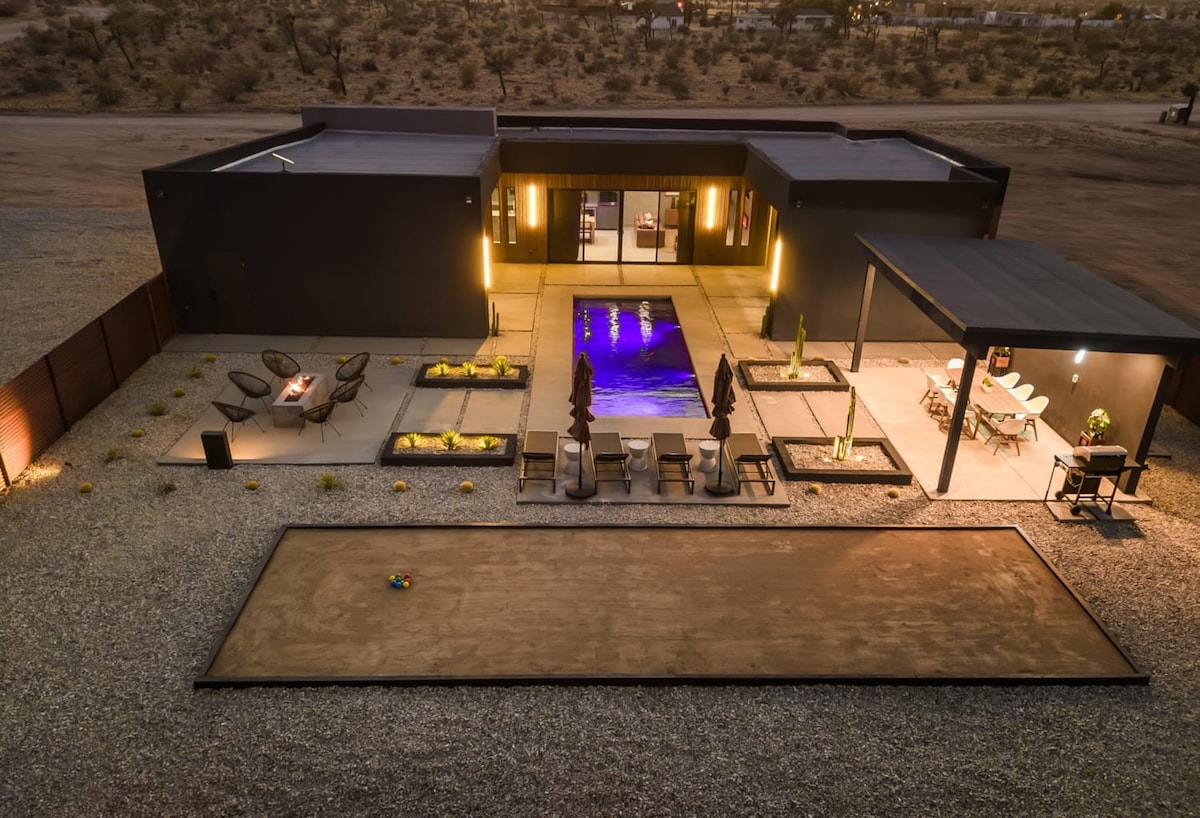 An aerial view captures the modern layout of the property, featuring a rectangular pool at its center, surrounded by lounge chairs and a dining area. A bocce ball court is seen in the foreground, while desert landscaping enhances the serene atmosphere.