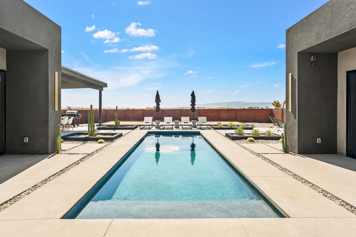 A luxury swimming pool is framed by modern structures, showcasing clear water under the bright blue sky. Loungers are positioned along the pool's edge, with shaded umbrellas providing respite from the sun. Surrounding desert landscaping adds to the tranquil ambiance.