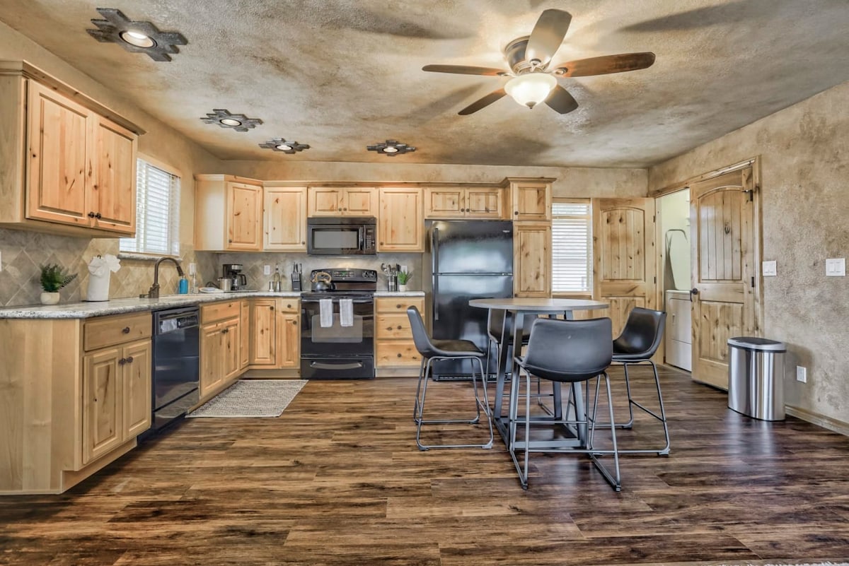 A bright and spacious kitchen features wooden cabinetry and stainless-steel appliances. A dining table for four is positioned center, surrounded by black chairs. A ceiling fan provides additional comfort, and natural light pours in through the window above the sink.
