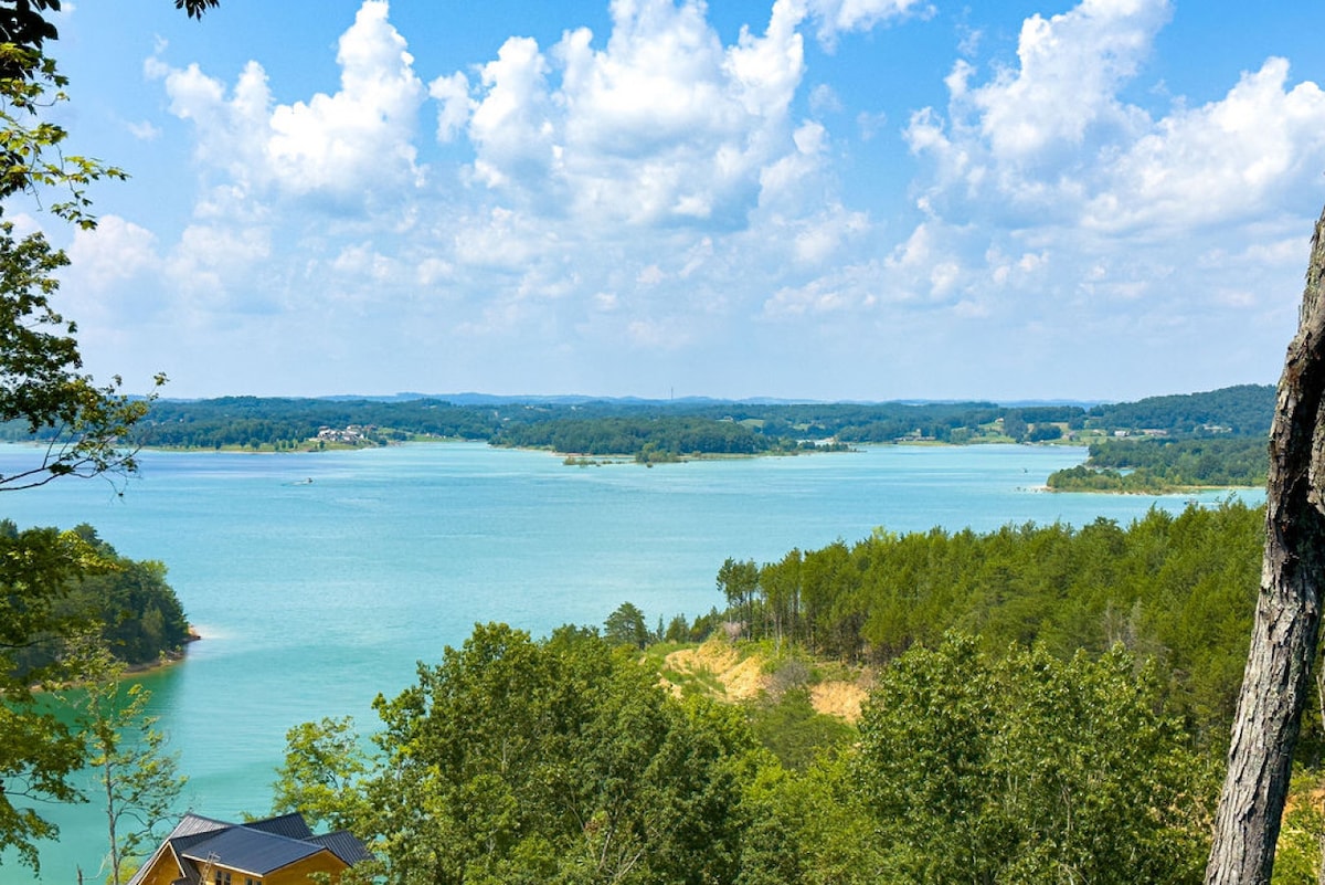 A panoramic view of Douglas Lake is featured, showcasing its clear turquoise waters surrounded by lush greenery and rolling hills. The sky is filled with scattered clouds, providing a serene backdrop to the landscape. Visible hills in the distance create a sense of depth.