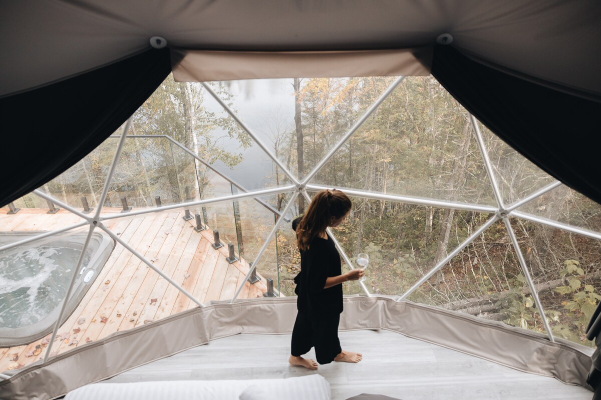 A spacious dome interior features a child standing barefoot on wood flooring, overlooking a serene view through large, rounded windows. Outside, a hot tub and wooden deck are visible, surrounded by trees and natural scenery.
