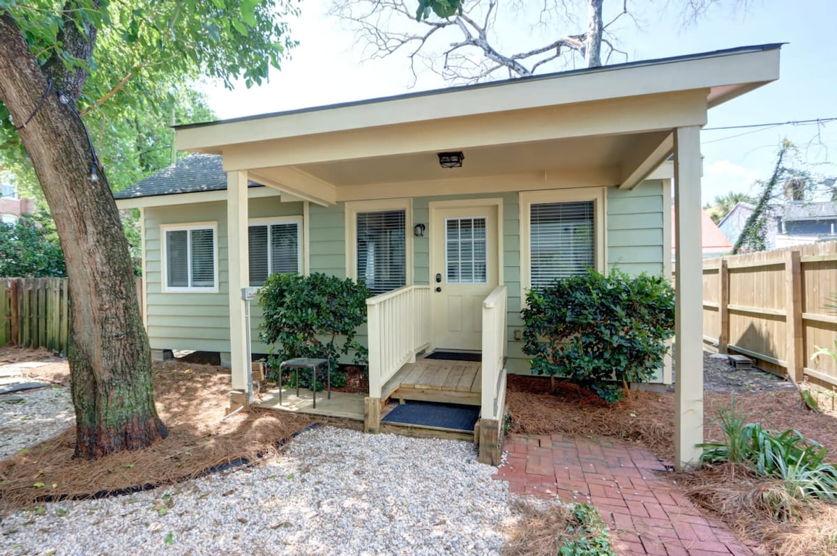 The exterior of the cottage is shown, featuring a welcoming porch with steps leading to a small wooden deck. Green siding and well-maintained shrubbery surround the entrance, while a mixture of gravel and brick pathways are present in the yard.