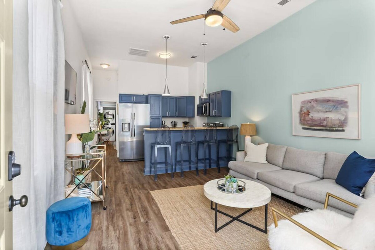 The living area features a light-colored sectional sofa paired with a circular marble coffee table. A kitchen with navy cabinets is visible in the background, along with four bar stools. Natural light streams through the windows, illuminating the room's soothing palette.
