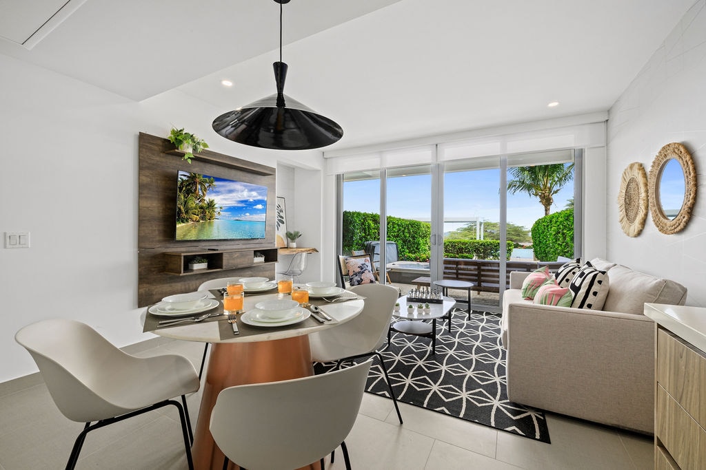 A modern living and dining area is displayed, featuring a round table set for dining, complemented by light-colored chairs. A wall-mounted flat-screen TV is positioned above a stylish wooden shelving unit. Large windows offer views of an outdoor seating area lush with greenery.
