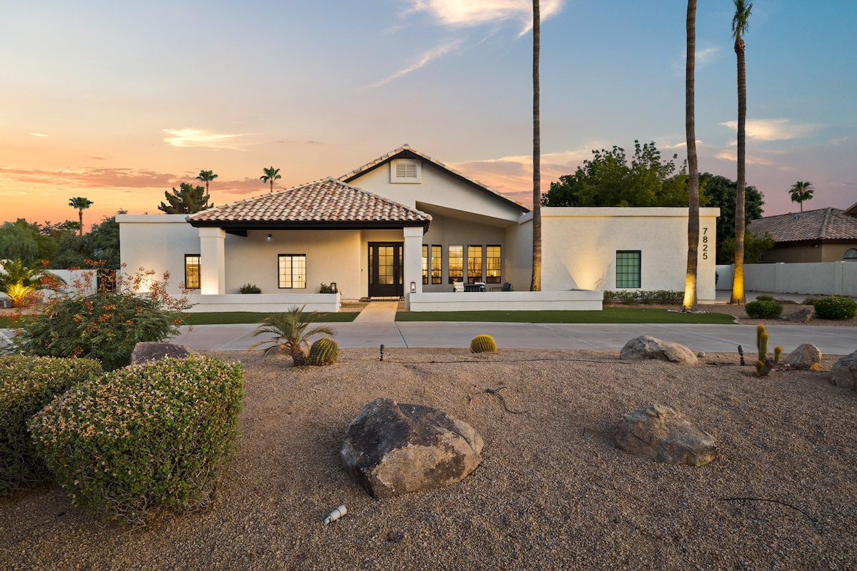The home is showcased against a serene sunset, featuring a welcoming facade and a tile roof. Lush shrubs and large rocks frame the entrance, enhancing the spacious landscape while tall palm trees rise nearby, contributing to the inviting atmosphere.