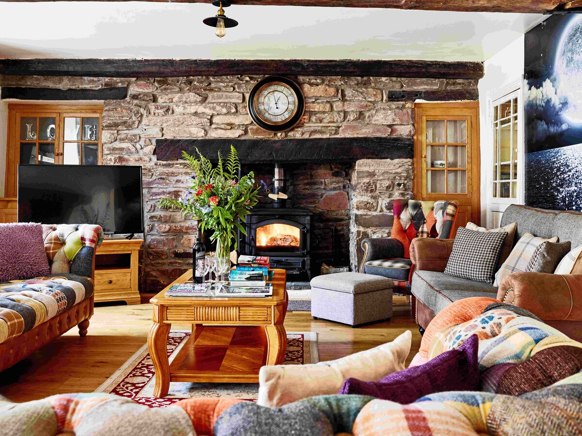 A cosy lounge area features a wood-burning stove set within a traditional stone fireplace. Comfortable seating options, including a large sectional sofa and armchairs, are arranged around a wooden coffee table. A television is positioned nearby, complemented by decorative plants and a vibrant display of cushions.