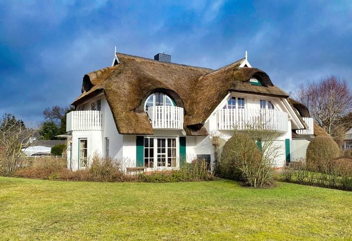 A charming thatched-roof house is surrounded by a well-kept garden. Multiple balconies with white railings add character to the exterior, while lush greenery stretches across the lawn. A bright sky complements the tranquil atmosphere of the residence.