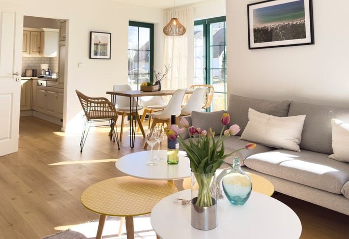 A bright living area is depicted, featuring a light gray sofa complemented by decorative cushions. In the foreground, a coffee table holds a vase of flowers. A dining table with white chairs is visible in the background, along with a glimpse of the kitchen area through an open doorway.