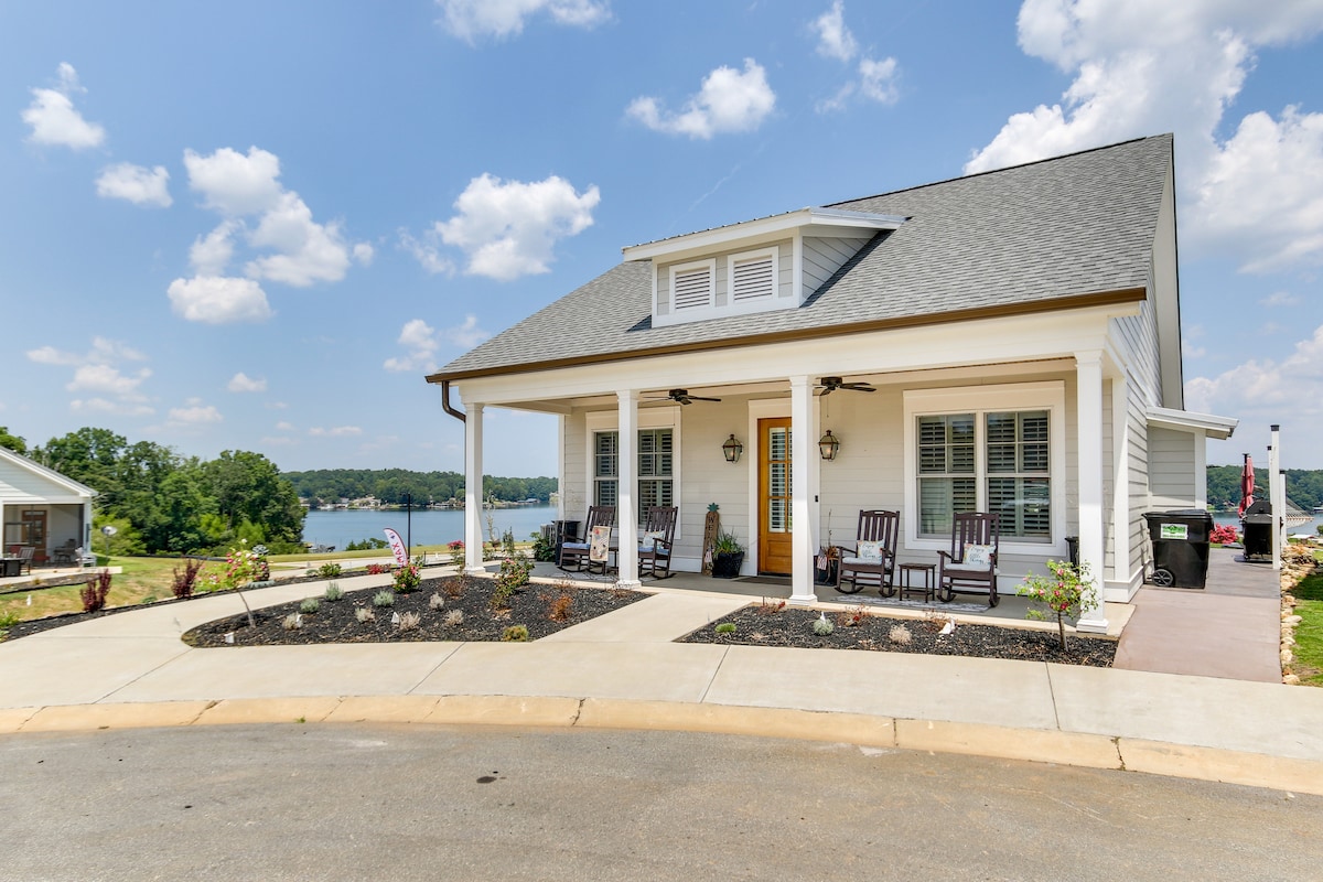 A charming cottage exterior is shown, featuring a covered porch with rocking chairs. Landscaped flower beds are visible, enhancing the inviting entryway. The backdrop showcases a serene lake beneath a partly cloudy sky, contributing to the peaceful ambiance of the property.