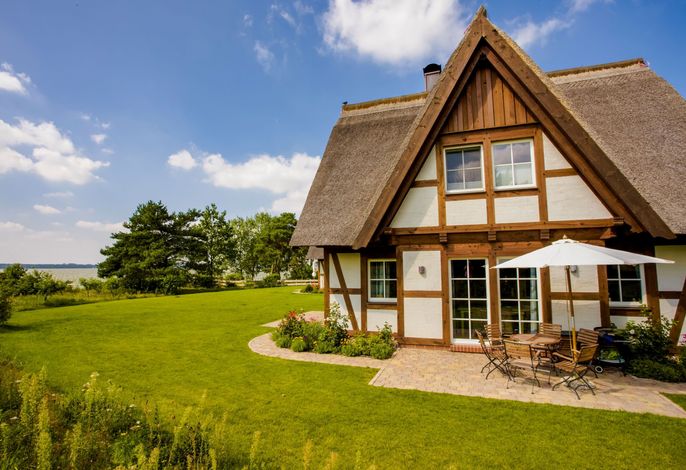 The half-timbered house is surrounded by lush greenery, with a neatly landscaped lawn in front. A terrace features a dining set under a shaded umbrella, offering a scenic view of the nearby water and sky. The thatched roof complements the house's rustic charm.