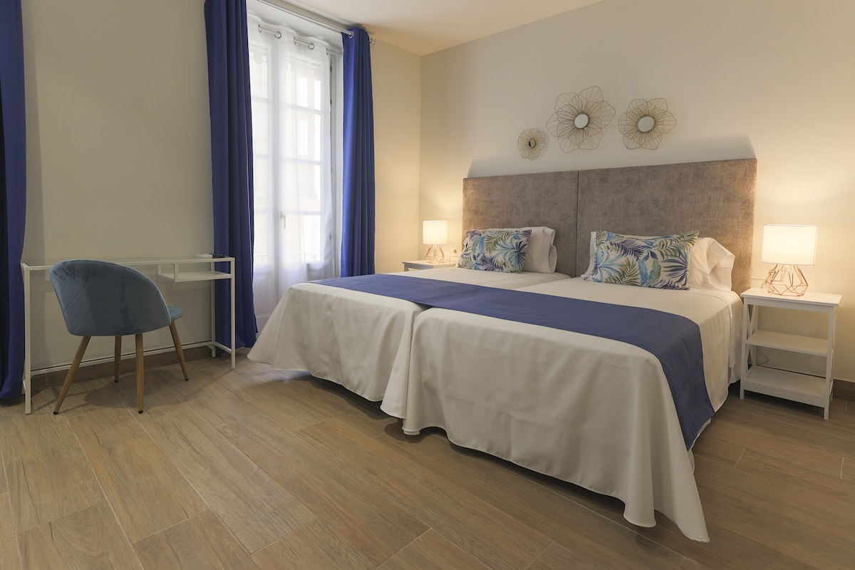 A spacious double room features two well-appointed beds with white linens and decorative blue throws. Two bedside tables with lamps are positioned on either side of the beds. A desk and chair are placed against a wall, and large windows allow natural light to fill the room.