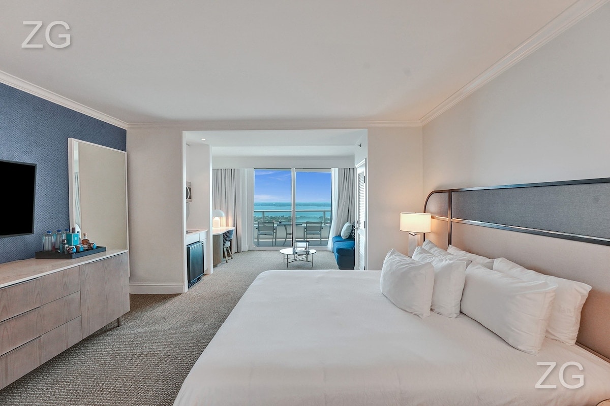 A spacious studio is highlighted by a king-size bed adorned with crisp white linens. A comfortable seating area is visible near large glass doors, opening to a balcony that offers expansive ocean views. Soft natural light filters through sheer curtains.