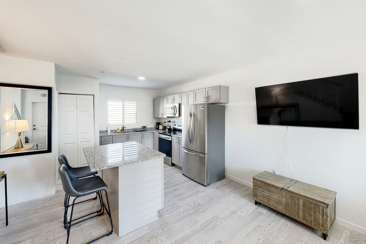 An open-concept kitchen and living area is displayed, featuring sleek stainless steel appliances and a large granite island with seating for four. A wall-mounted television and a wooden coffee table provide functional elements to the space, illuminated by natural light from nearby windows.