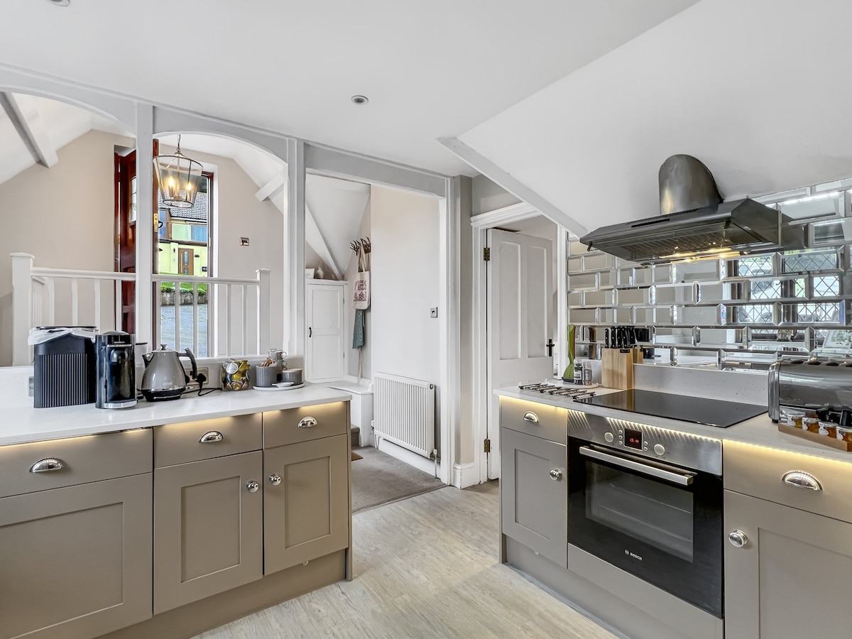 A modern kitchen is presented with a spacious layout, featuring a sleek stovetop and oven along with various kitchen appliances. Light reflects off the mirrored backsplash, enhancing the bright atmosphere. The adjoining entrance area can be glimpsed in the background, adding depth to the scene.