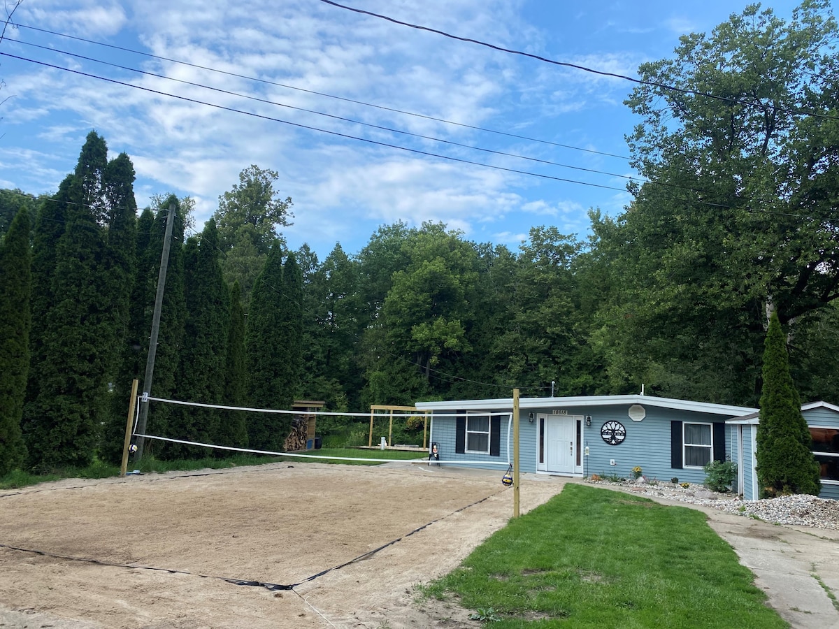 A private beach volleyball court is set in a sandy area, surrounded by tall green hedges and trees, offering a sense of seclusion. The spacious lawn is well-maintained, with the rental property visible in the background featuring a circular wall clock and an inviting porch.
