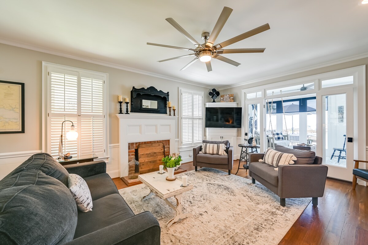 A well-appointed living area features comfortable seating with dark gray sofas and a soft area rug. A wooden coffee table sits in the center, while a large ceiling fan adds a touch of sophistication. Natural light streams through the windows, illuminating the space.