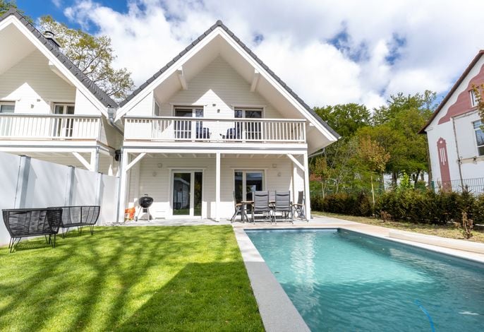 A two-story house features a light-colored exterior with a spacious balcony. A well-maintained lawn surrounds a private pool, and patio furniture is arranged neatly nearby. Sunlight beams down, highlighting the inviting outdoor space perfect for relaxation.