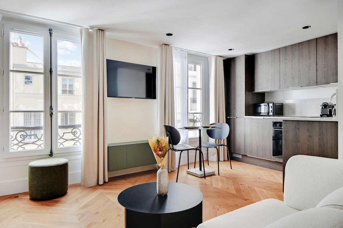 A modern living space features large windows that allow natural light to brighten the area. A wall-mounted television is positioned above a low coffee table. A small dining table with two chairs is set near an open kitchen, showcasing wood cabinetry and appliances.