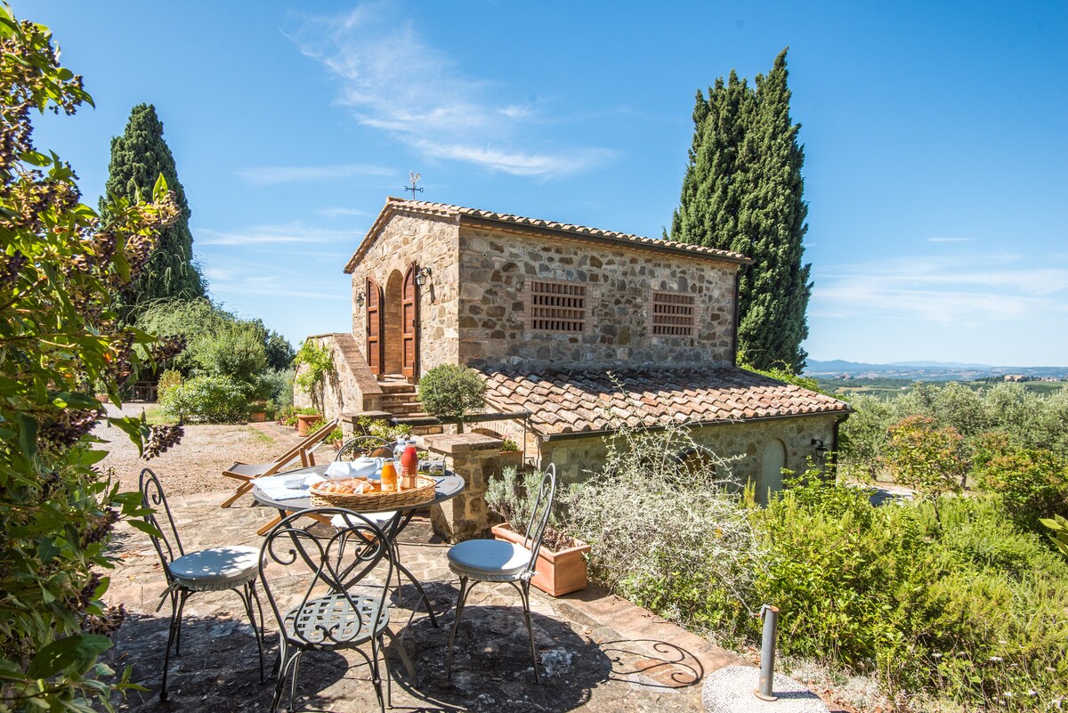 The farmhouse showcases a rustic stone exterior surrounded by greenery. A small outdoor dining area with a round table and chairs is set in front, accompanied by a platter of refreshments. Sunlight gently illuminates the scene, creating a serene ambiance.