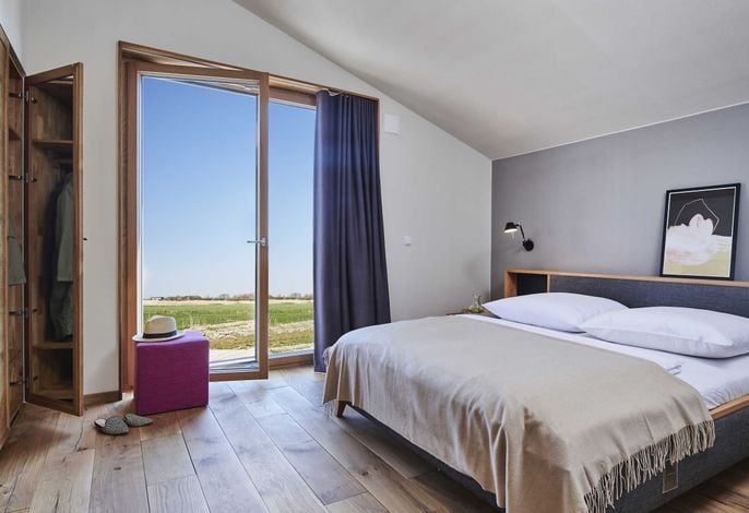 A serene bedroom features a spacious bed dressed in soft linens, with a throw blanket neatly arranged. Natural light floods in through large glass doors offering views of the outdoors. A wooden wardrobe stands to the side, complemented by a simple purple stool and a hat.