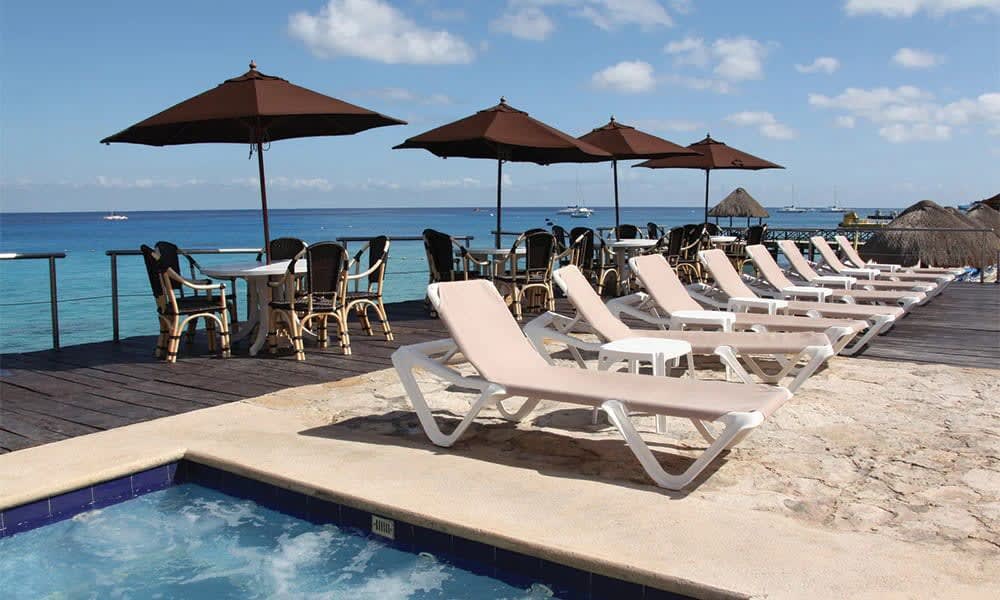 Lounge chairs are arranged in a row adjacent to the water, under large umbrellas. Tables and chairs are set nearby, offering a view of the calm sea. A hot tub is located at the foreground, with inviting water reflecting the sky.