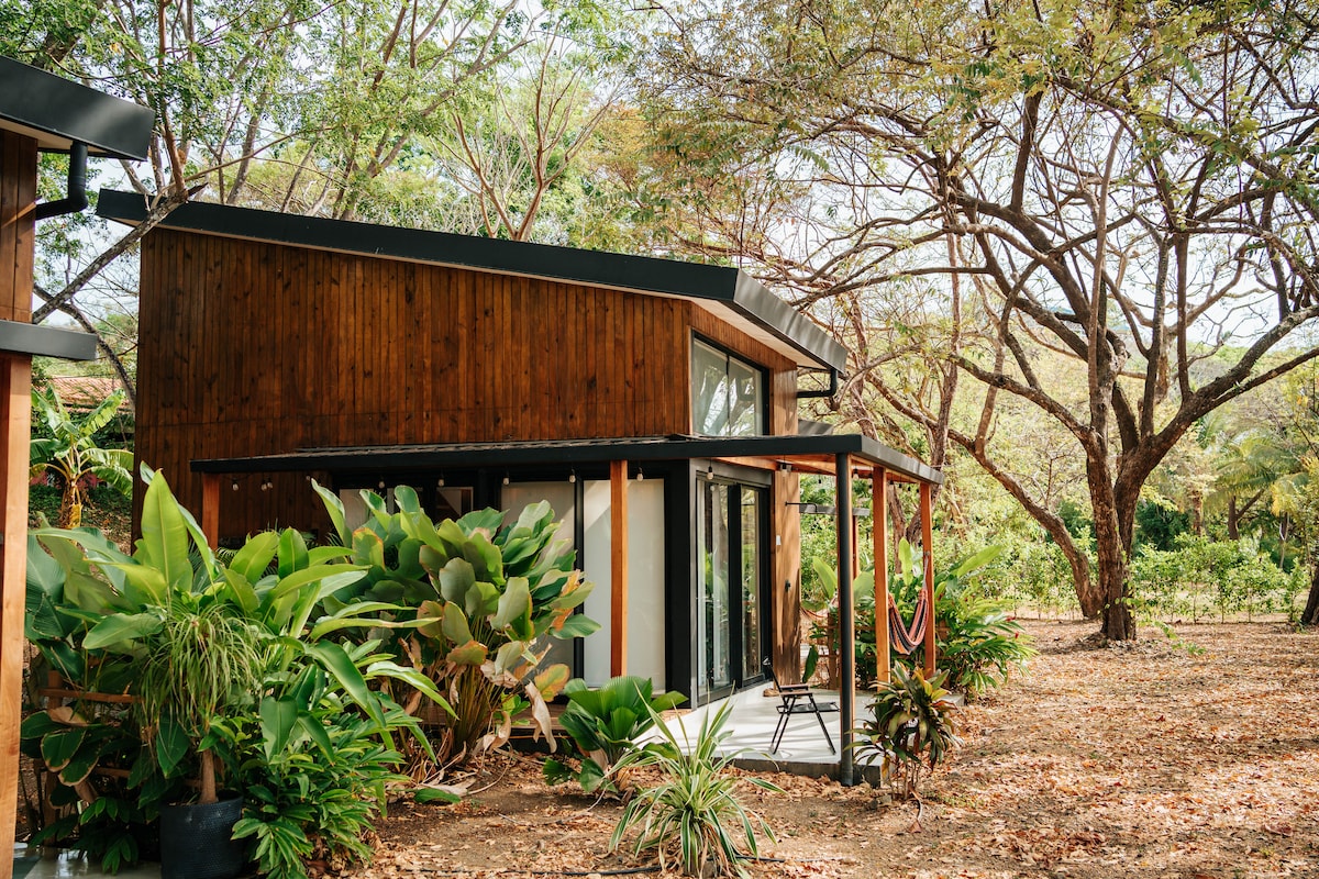 A modern tiny house with wooden accents is set amidst lush greenery. Large glass doors create a seamless connection to the outdoors, while tropical plants surround the porch area. The natural landscape enhances the overall minimalist design of the structure.