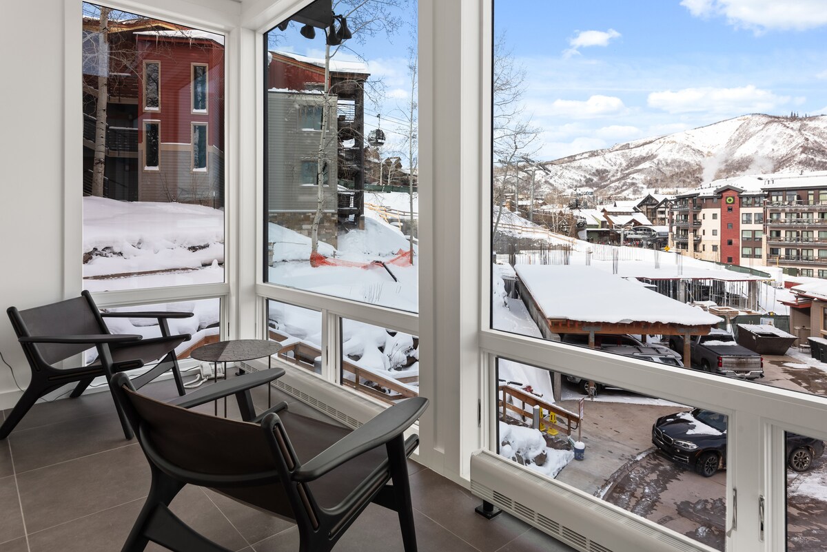 A bright corner seating area features two modern chairs and a small table, offering views of snow-covered mountains and nearby buildings through expansive windows. The floor is tiled, enhancing the open and airy feel of the space.