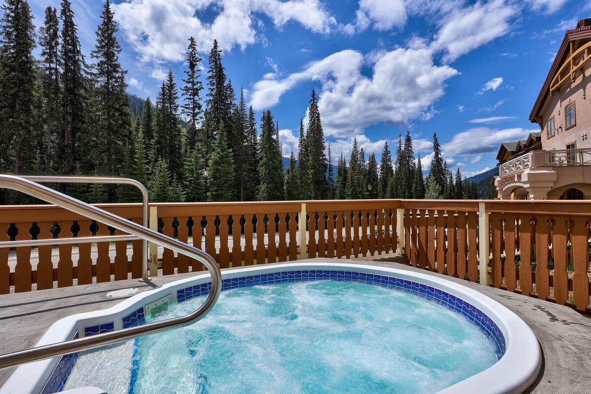 A hot tub is positioned on a terrace, surrounded by wooden fencing. The view showcases tall evergreen trees against a bright blue sky with scattered clouds. The water in the hot tub appears inviting and bubbly, suggesting a relaxing atmosphere.