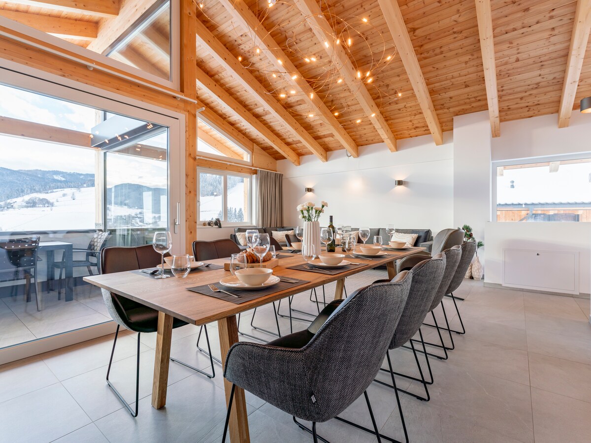 A spacious dining area features a large wooden table surrounded by gray chairs. The high ceiling with wooden beams is complemented by modern lighting. Large glass doors open to a terrace, allowing natural light and scenic mountain views to enhance the space.