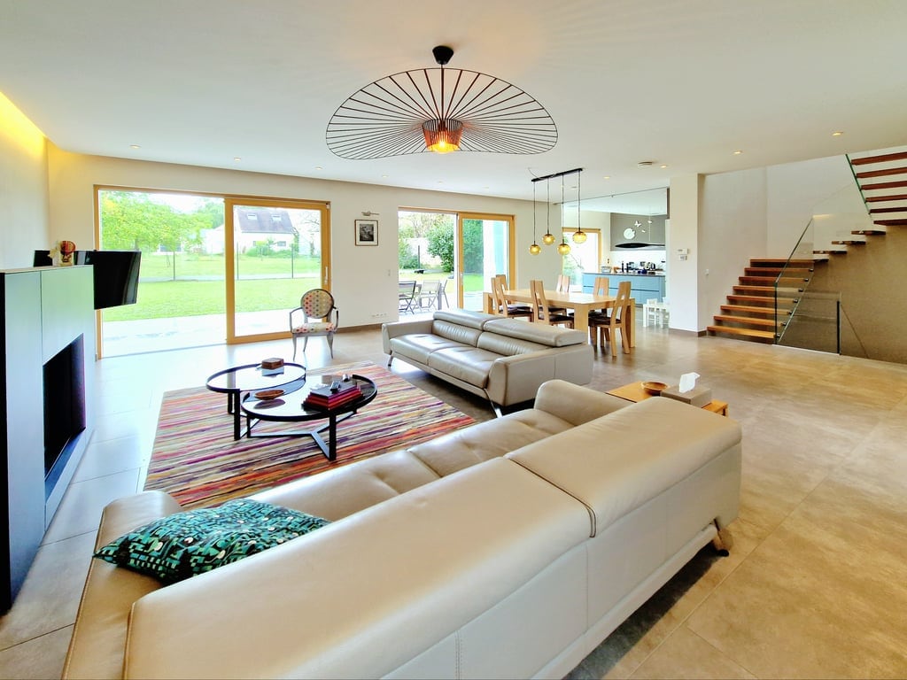 A spacious living area features a modern design with large glass doors that open to the garden. Natural light floods the space, highlighting the comfortable seating and dining area. A stylish spiral staircase adds a contemporary touch to the overall aesthetic.