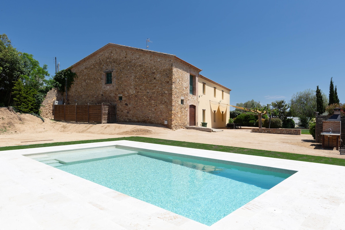 The villa is shown with its rustic stone exterior and large windows, set against a clear blue sky. A swimming pool is situated in the foreground, featuring crystal-clear water and surrounded by a well-maintained lawn, enhancing the outdoor space.