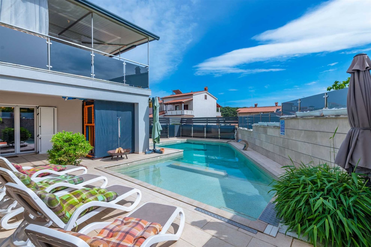 A clear view of a private heated pool surrounded by sunbeds and parasols. The pool area features a well-maintained patio, while the surrounding greenery adds a touch of nature. The sky is bright and blue, enhancing the outdoor relaxation space.