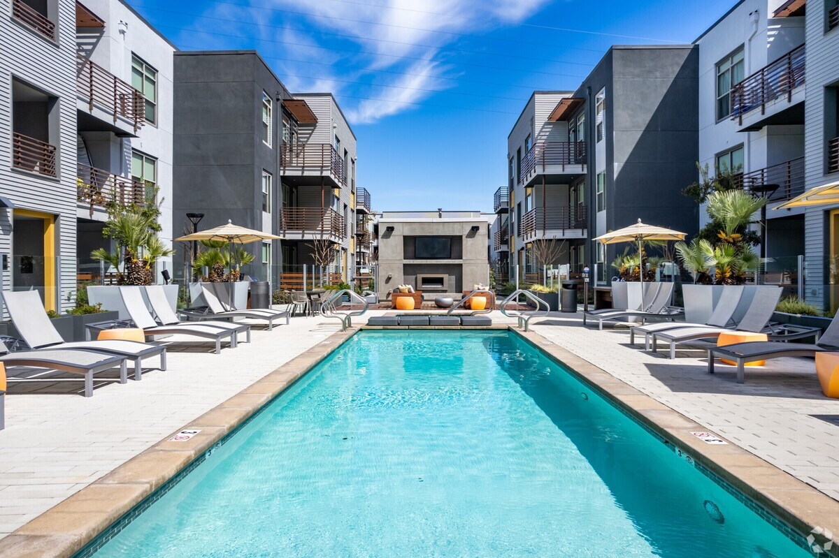 The resort-inspired pool is surrounded by lounge chairs and umbrellas, creating a relaxing atmosphere. Two modern apartment buildings frame the space, with a fire pit visible in the background. Clear blue skies reflect on the water's surface, enhancing the inviting setting.