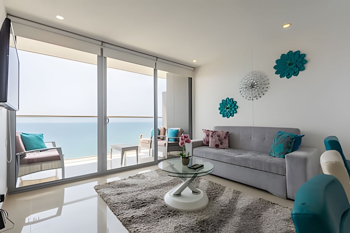 A modern living area is highlighted by large glass doors that open to a stunning ocean view. Light-colored furniture complements the space, while decorative blue accents and a plush gray sofa add a pop of color. A round glass table rests atop a soft area rug beside an indoor plant.