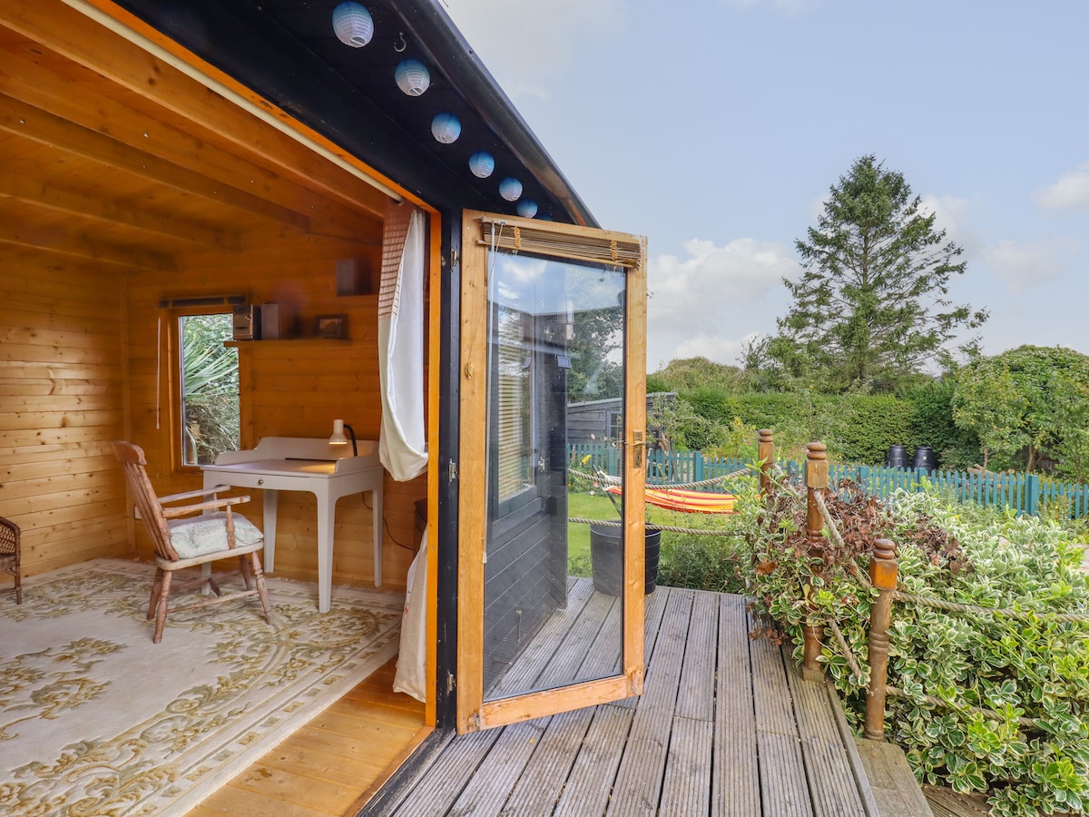 The image captures a wooden structure with large glass doors opened to a deck area. Inside, a simple desk is present, accompanied by a wicker chair. Lush greenery is visible outside, along with a glimpse of the garden and additional outdoor features.