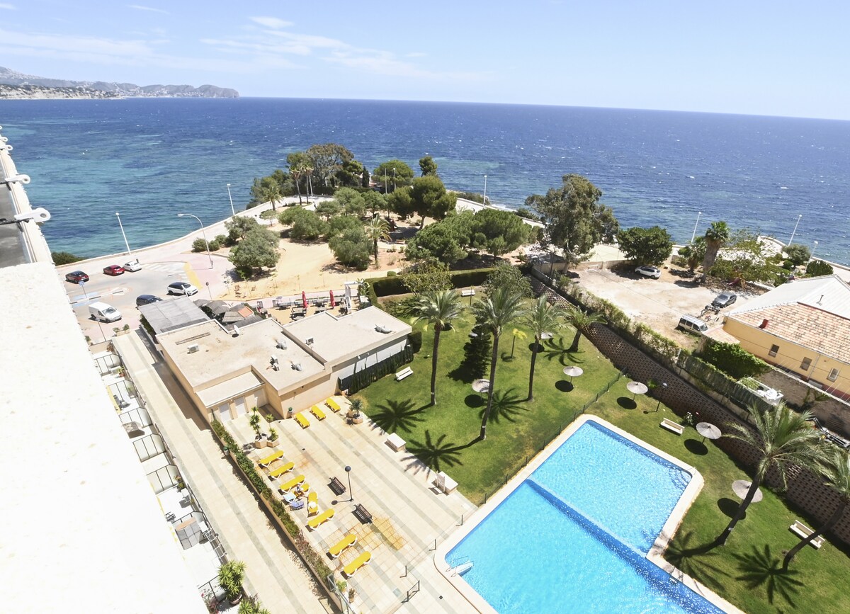 An aerial view captures the communal swimming pool surrounded by well-maintained grass and palm trees. The blue waters of the sea stretch toward the horizon, with a sandy area and pathways visible in the foreground. The landscape reflects a coastal setting inviting relaxation.
