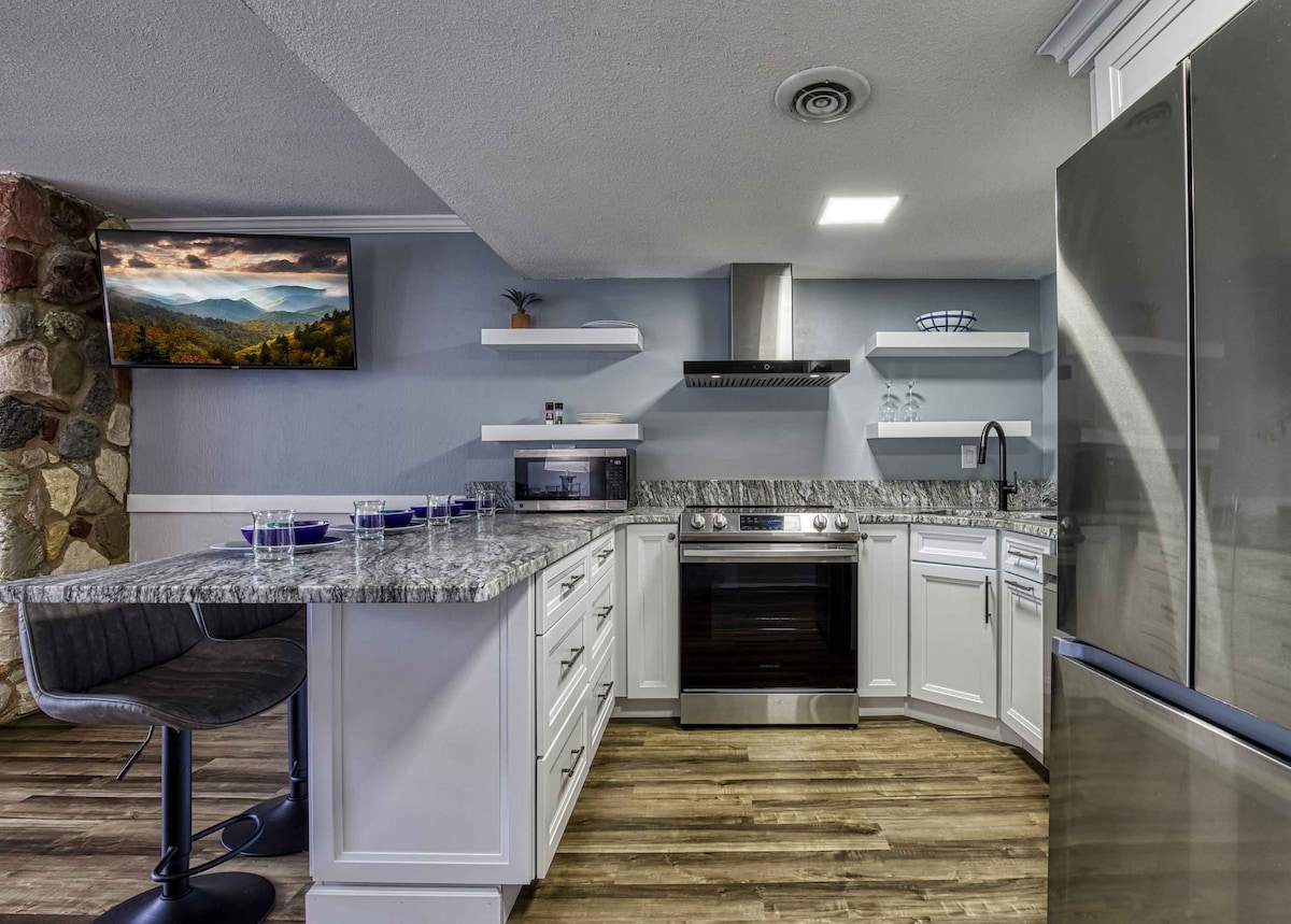 The modern kitchen features polished countertops and stainless steel appliances, including an oven and microwave. Open shelving displays tasteful decor, while a large television mounted on the wall adds convenience. A kitchen island with bar seating invites casual dining and gathering.