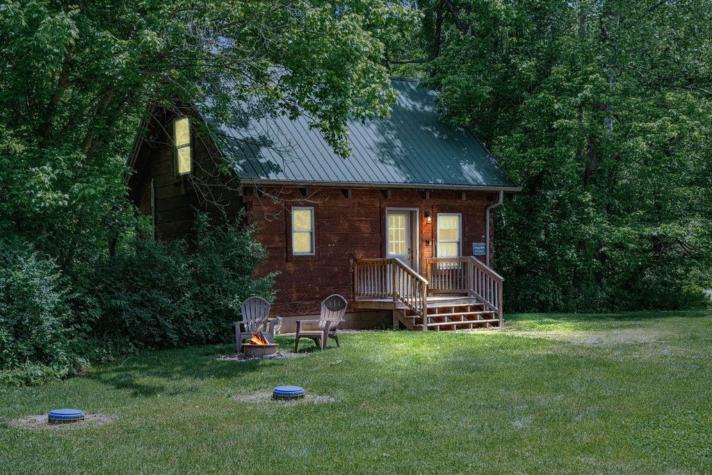 A rustic log cabin is framed by lush greenery, featuring a welcoming front porch with steps leading up to two doors. Two wooden chairs sit near a small fire pit on the manicured lawn, inviting relaxation in nature.