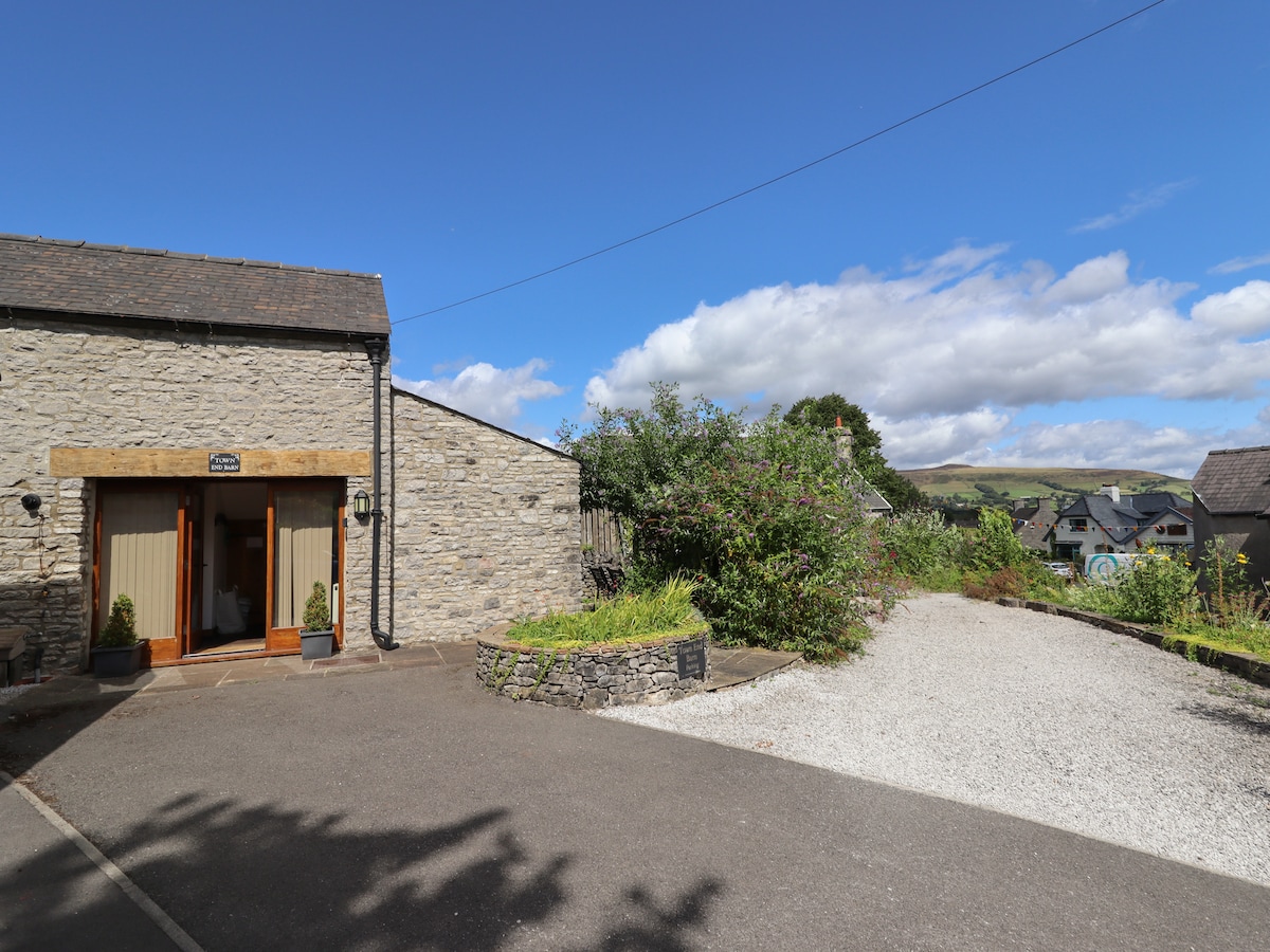 A stone exterior of Town End Barn is highlighted against a clear blue sky. The entrance features double doors framed by plants, with a gravel driveway leading in. Surrounding greenery and hills in the distance enhance the rural setting.