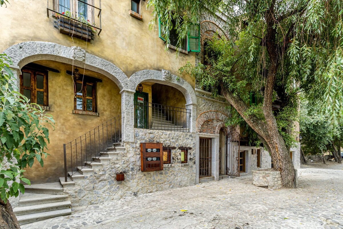 The exterior of a charming building is presented, featuring stone walls and archways. A staircase leads to the entrance, with vibrant green shutters adorning the windows. Lush trees provide shade around the area, enhancing the inviting and rustic appeal of the environment.