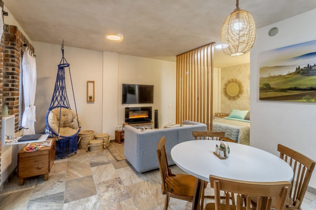 A cozy living area features a comfortable sofa and a circular dining table for four, complemented by wooden dining chairs. A smart TV is mounted on the wall, while a stylish lamp hangs from the ceiling. Natural light enters through large windows, enhancing the serene ambiance.