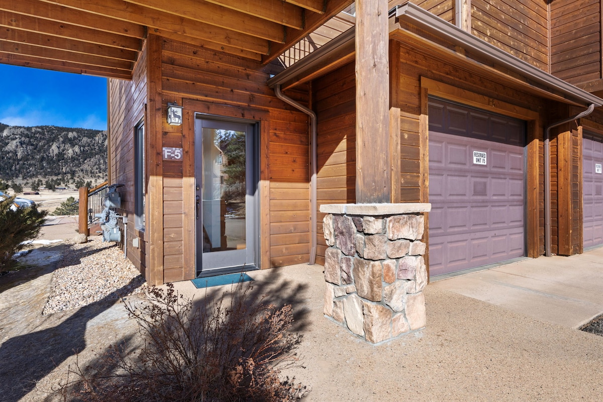 The entrance to the two-bedroom condo features a wooden exterior with natural stone accents. A pathway leads to the door, framed by shrubs and gravel. The garage door is visible, blending seamlessly with the rustic architecture.
