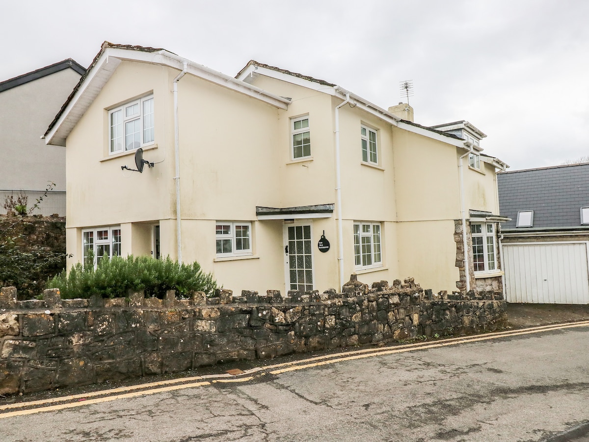 A detached cottage is viewed from the street, displaying a light-colored exterior with multiple windows. A stone wall borders the property, and a small garden area is visible at the front. Rooflines vary, contributing to the unique architecture.