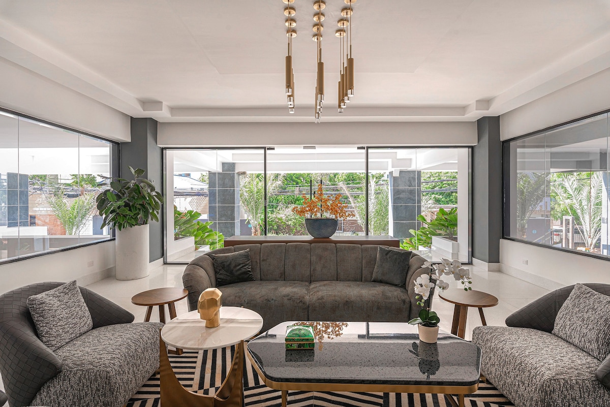 The spacious living area features a comfortable sectional sofa surrounded by armchairs. Natural light floods in from the large windows, highlighting indoor plants and a decorative centerpiece on the coffee table. A modern chandelier adds a touch of elegance to the space.