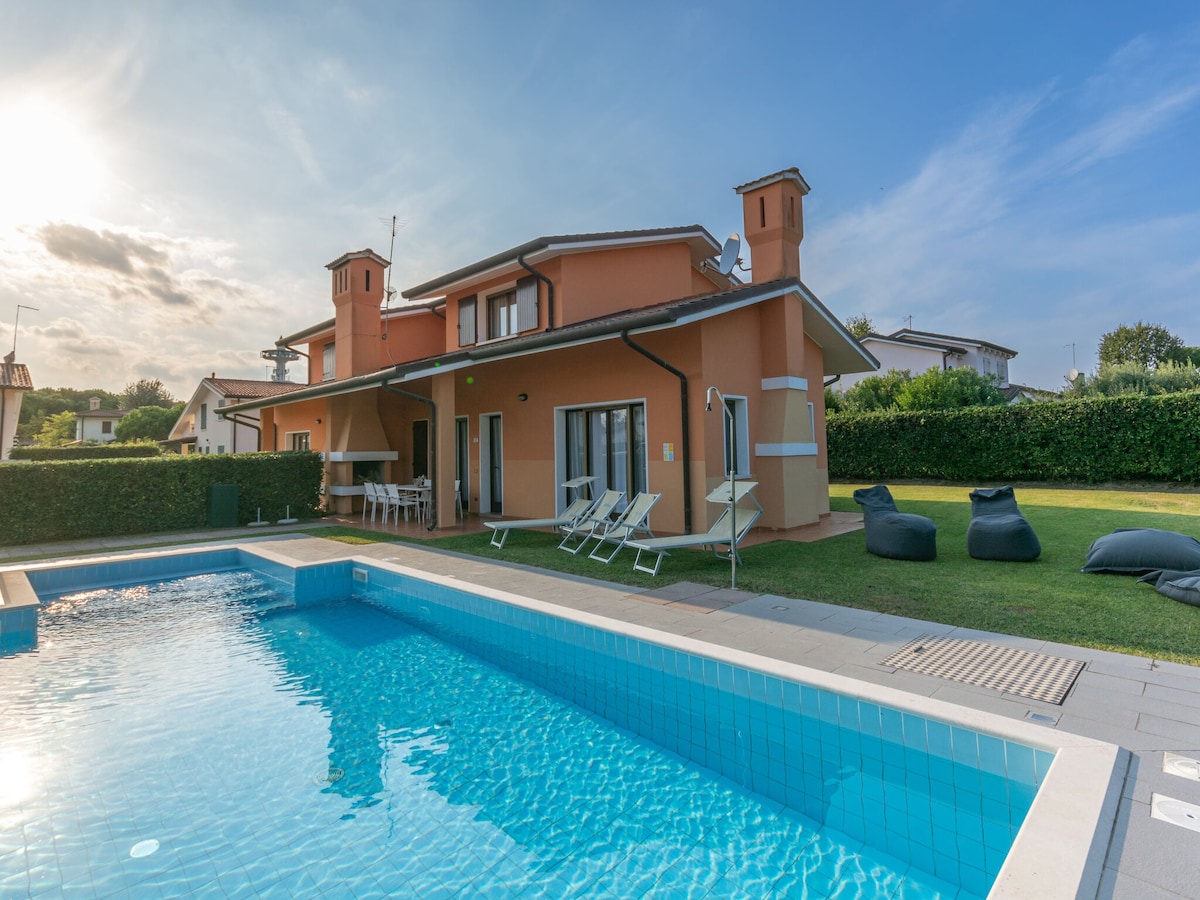 A modern villa is depicted with a spacious private pool reflecting the blue sky. The exterior showcases a blend of orange and white, complemented by two chimneys. Garden furniture including loungers and chairs is arranged beside the pool, surrounded by well-maintained greenery.