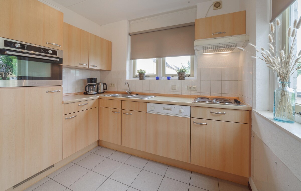 A functional kitchen is presented, featuring wooden cabinetry and modern appliances. A microwave and coffee maker are visible on the countertop. Natural light enters through a window adorned with light shades, while a vase of decorative grass adds a touch of greenery.