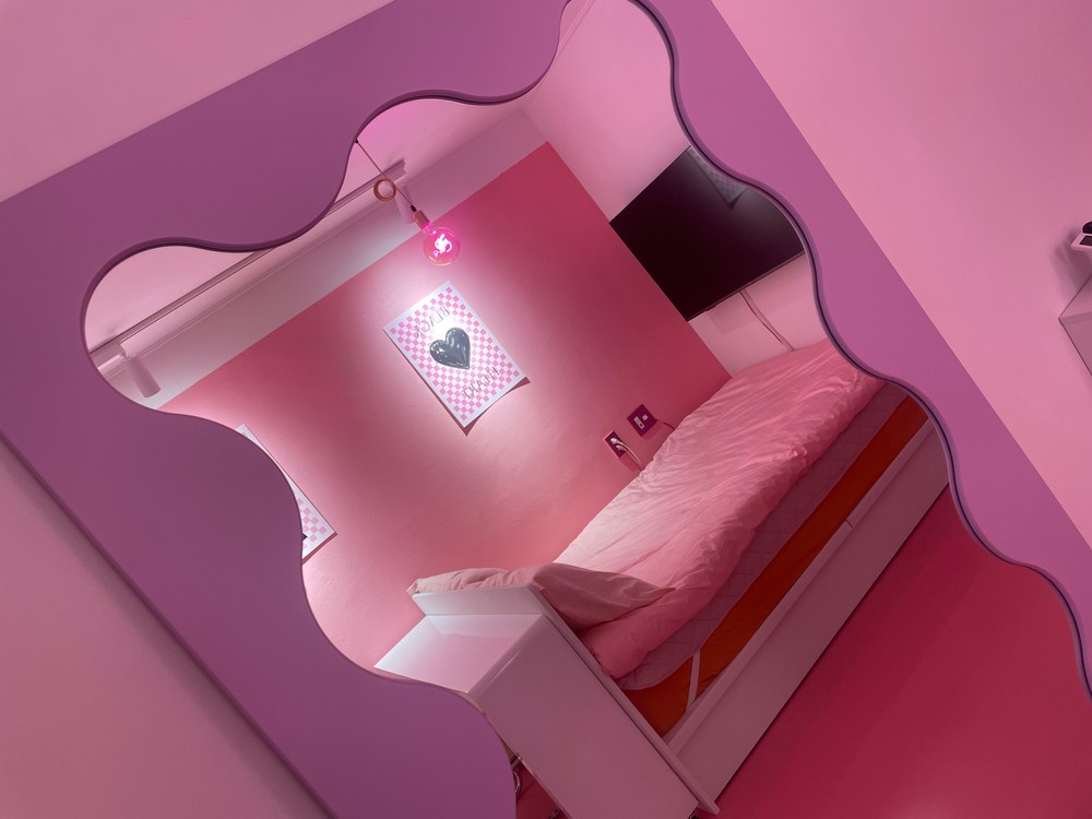 A spacious bedroom with a soft pink color scheme is displayed. The room features a large bed dressed in light bedding. A decorative mirror with a wavy edge reflects the area, while a modern lighting fixture casts a gentle glow. Artwork is visible on the wall.