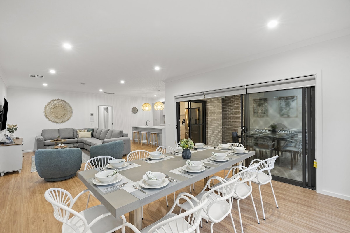 A spacious dining area is showcased with a long table set for a meal, featuring decorative plates and glassware. Soft lighting enhances the room, which is connected to a cozy living space with a sectional sofa, leading to glass doors opening to the outdoor area.
