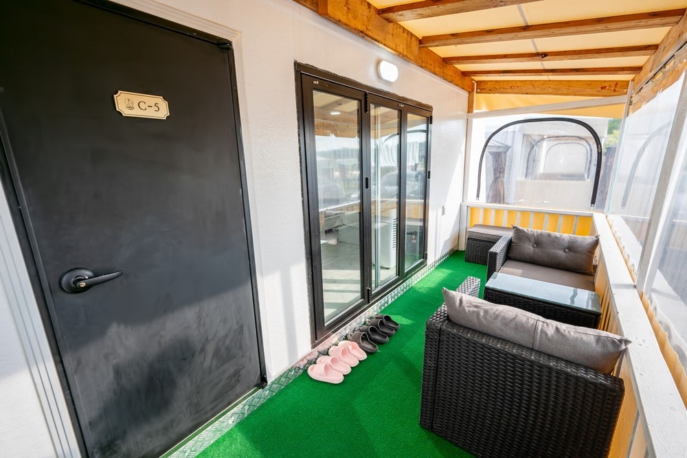 A welcoming terrace area features a black door with a clearly labeled sign. Two comfortable seating arrangements are provided, alongside a green carpeted floor. Multiple pairs of slippers are neatly placed at the door, enhancing the inviting entryway.